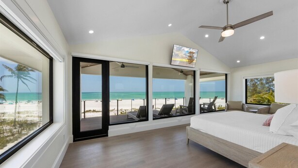 A bedroom with a large bed, a ceiling fan, and a view of the beach through large windows. - True Beachfront!  2 Beachfront Masters - Best Location!! (Anna Maria Island)