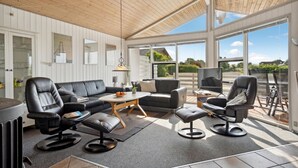 Una sala de estar moderna con techo de madera, ventanas amplias, un sofá de cuero negro, dos sillones, una mesa de centro y un televisor.