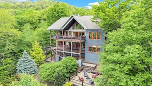 A multi-story cabin with a balcony and a deck, surrounded by dense forest.
