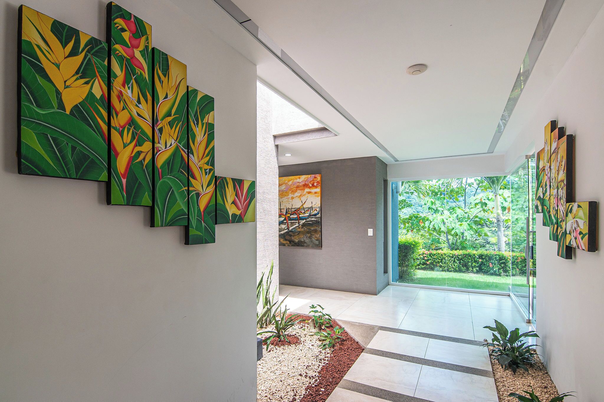 A hallway with a series of paintings on the walls, a glass door leading to a garden, and a tiled floor.