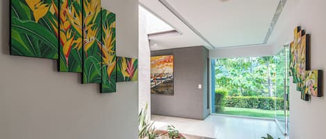 A hallway with a series of paintings on the walls, a glass door leading to a garden, and a tiled floor.