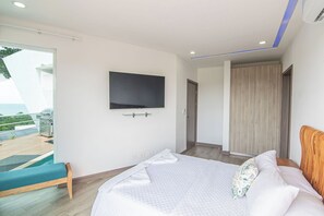 A bedroom with a bed, a TV mounted on the wall, a wooden wardrobe, and a balcony with a view of the sea.