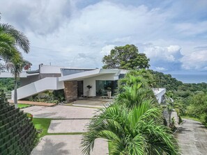 Een modern huis met een schuin dak, omgeven door weelderige begroeiing en uitzicht op zee.