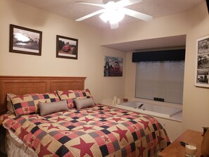 A bedroom with a large bed, a ceiling fan, a window with blinds, and a small table with a candle.