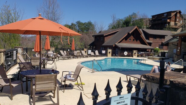 A pool area with lounge chairs and umbrellas, a sign indicating social distancing, and a wooden building in the background. - Mountain Time in Pigeon Forge Close to Dollywood (Pigeon Forge)