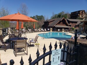 A pool area with lounge chairs and umbrellas, a sign indicating social distancing, and a wooden building in the background.