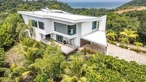 A modern white house with large windows, surrounded by lush greenery and overlooking a coastal view. - Luxury 4BR Ocean View with Infinity Pool & Jacuzzi (Playa Hermosa)