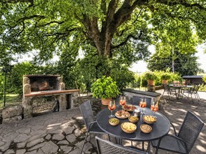An outdoor patio with stone flooring, a round table with chairs, a grill, and potted plants. - La Quercia by Interhome (Orte)