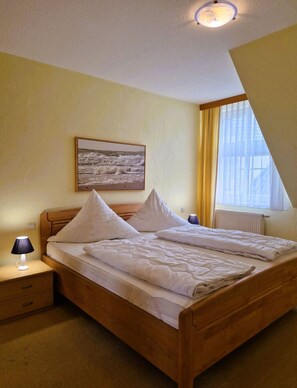 A bedroom with a wooden bed, white bedding, a nightstand with a lamp, a framed picture on the wall, and a window with curtains.