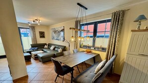Ein Wohnzimmer mit einer Couch, einem Esstisch und einer Bank. Ein Fenster mit Vorhängen und Ausblick.