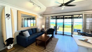 A modern living room with a sofa, coffee table, and a large mirror. There is a ceiling fan, a view of the ocean through large sliding doors, and a small table with a lamp. - Kiahuna Plantation #198 - The Best Beachfront Condominium on Kauai (Koloa)