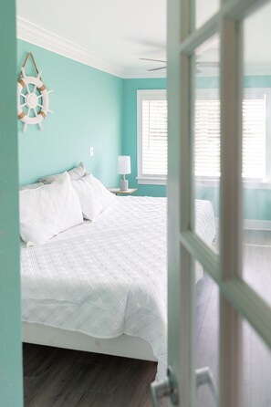 Une chambre à coucher comprenant un lit, une table de chevet, une décoration murale représentant un gouvernail de bateau et une fenêtre avec des stores.