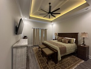 Un dormitorio con cama, mesitas de noche, un escritorio, un espejo, un ventilador de techo y una ventana con cortinas.