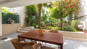 Une terrasse avec une table en bois et des chaises en osier, entourée d’une végétation luxuriante et de diverses plantes.