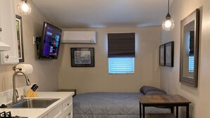 A compact bedroom with a bed, bedside table, and a small desk with a chair. A window with blinds is visible, and a mounted TV is on the wall. - Newly redone studio w/outdoor lawn & eating space (Azusa)