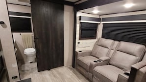 A compact RV interior with a toilet, a sofa, and a window with blinds.