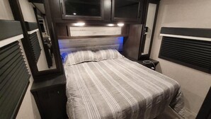 A compact RV bedroom with a bed, built-in TV, and overhead cabinets.