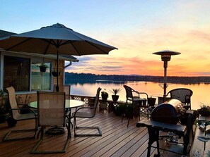 A deck with outdoor furniture, a grill, and a view of a lake at sunset.
