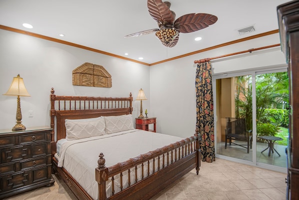 A bedroom with a wooden bed, two bedside tables, a ceiling fan, a decorative wall piece, and a view of a garden through a sliding glass door.