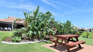 Een terras met een houten picknicktafel en banken, omgeven door weelderige tropische planten en een gebouw met een pannendak.