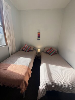 Une petite pièce bien éclairée, avec deux lits simples, une table de chevet avec une lampe et une décoration murale colorée.