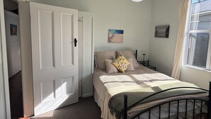 Un dormitorio con cama, puerta, ventana, lámpara de pared y una mesita.
