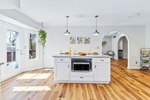 Een moderne keuken met witte kasten, een centraal kookeiland, hanglampen en uitzicht op buiten.