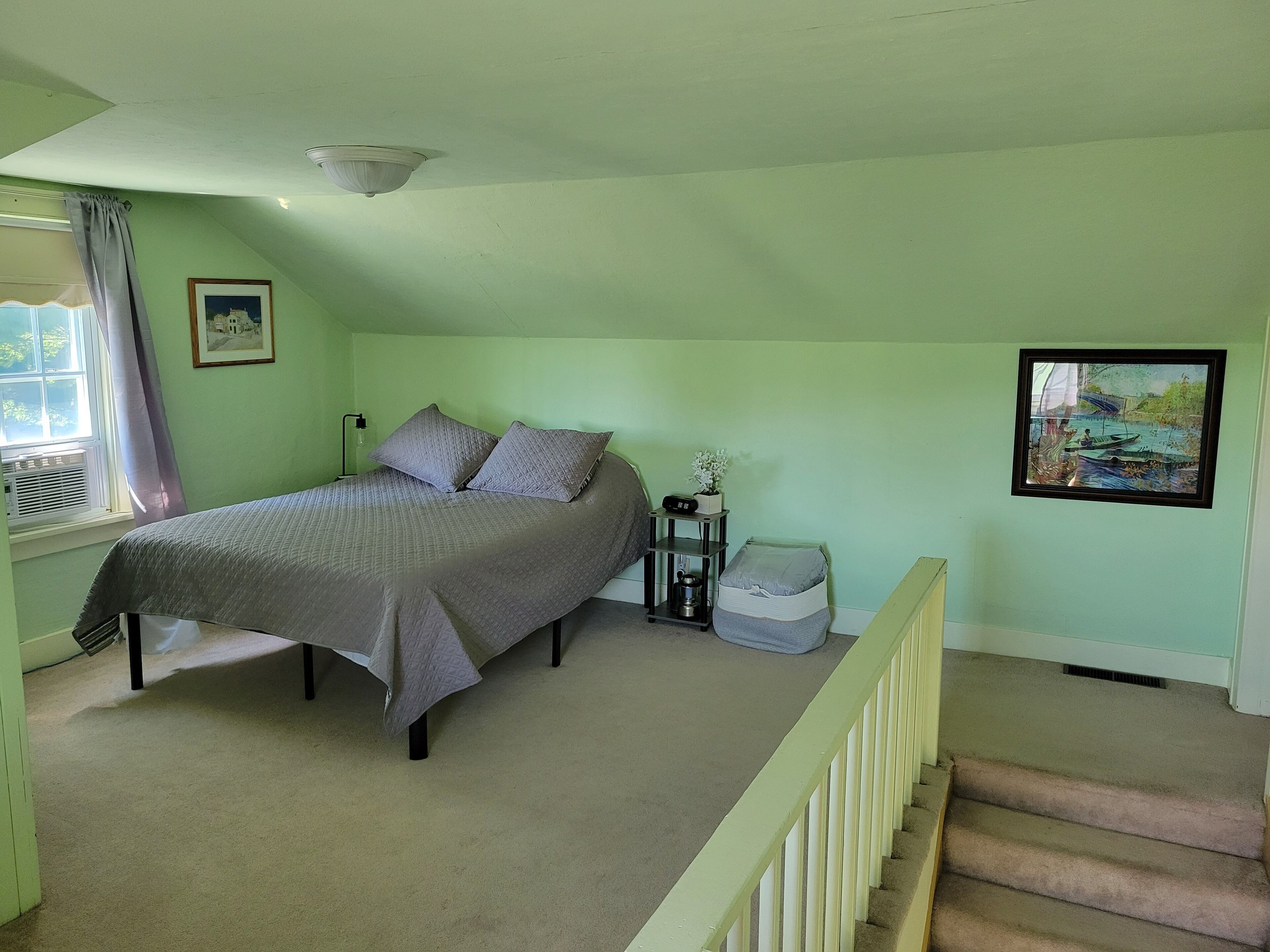 Ein Schlafzimmer mit einem Bett, einem Nachttisch, einem kleinen Tisch mit einer Pflanze, einem gerahmten Bild an der Wand und einer Treppe.