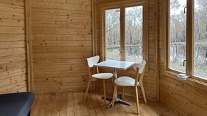 A wooden cabin interior with a dining table and chairs, large windows, and a view of a forest and water outside.