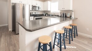 A modern kitchen with white cabinets, stainless steel appliances, a granite countertop, and three wooden bar stools.