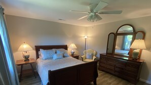 Une chambre à coucher comprenant un lit, une commode, une chaise, une lampe, un ventilateur de plafond et un miroir.