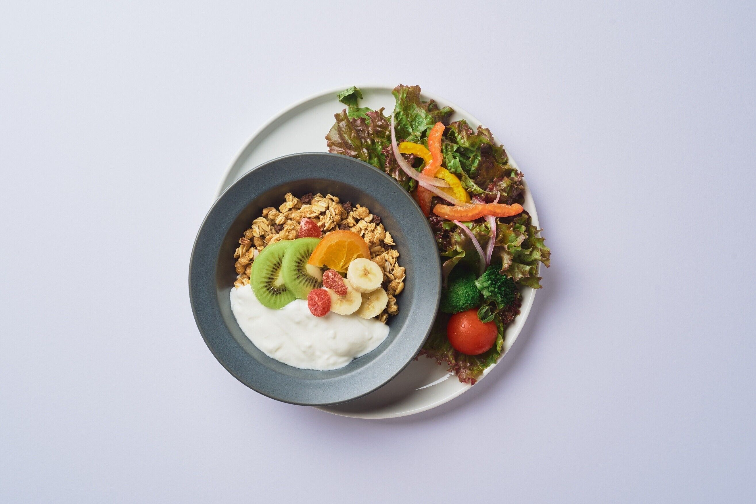 Un plato con un tazón de yogur cubierto de frutas y granola, y una ensalada con diversas verduras.