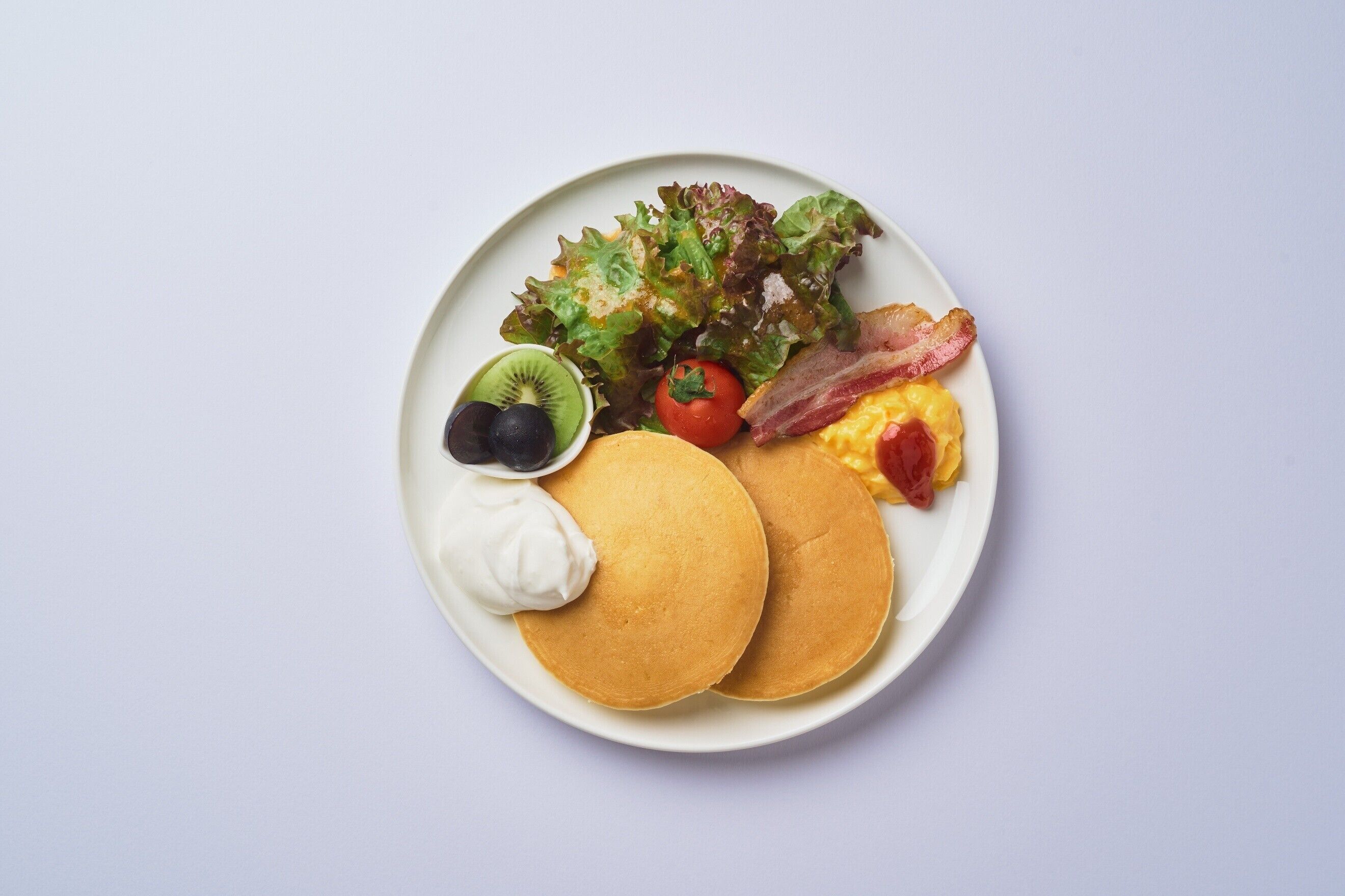 A plate with pancakes, bacon, eggs, a salad, and a slice of kiwi.