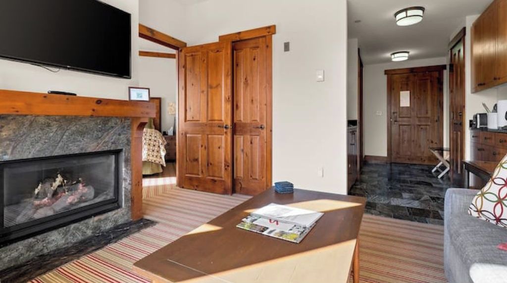 A living room with a fireplace, wooden doors, a television, and a kitchen area with cabinets.
