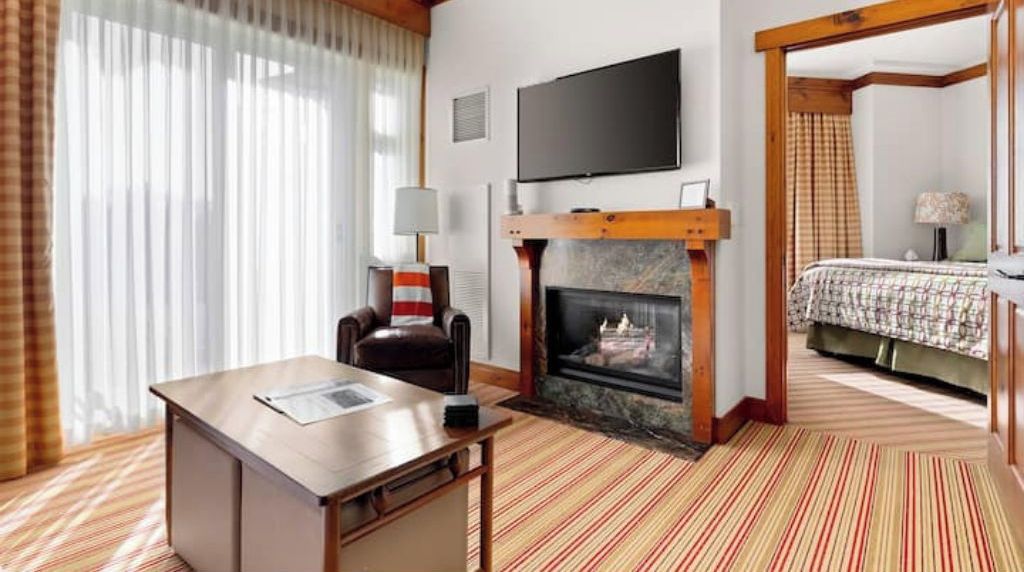 A hotel room with a desk, chair, fireplace, television, and a bed with a checkered bedspread.