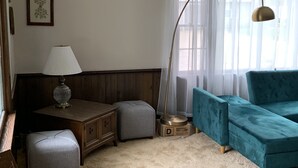 A living room with a teal sofa, a wooden side table with a lamp, a framed picture on the wall, and a large window with sheer curtains.