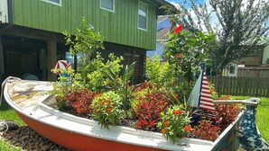 Una jardinera con forma de barco, con flores y una pequeña bandera estadounidense, ubicada en un patio cubierto de césped con una casa al fondo.