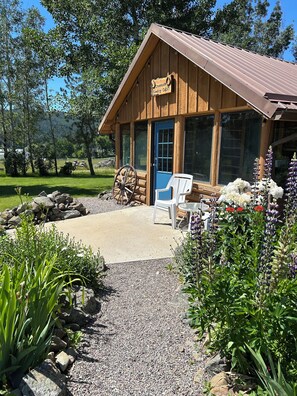 A wooden cabin with a blue door, a sign reading 'Creekside Cowboy Cabin,' and a gravel path leading to a white chair and table.
