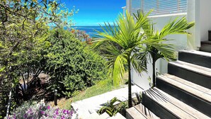 A view of a coastal area with a staircase leading to a building with white exterior and blue shutters, surrounded by lush greenery and a clear blue sky. - Villa NALA seaside pool standing, relaxation facing the lagoon feet in the water  (LE ROBERT)