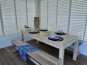 A dining area with a wooden table set for four, white blinds in the background, and a bench with a blue and white striped blanket. - Villa NALA by the Sea with a Pool: Relax with a View of the Lagoon, Feet in the Water (LE ROBERT)