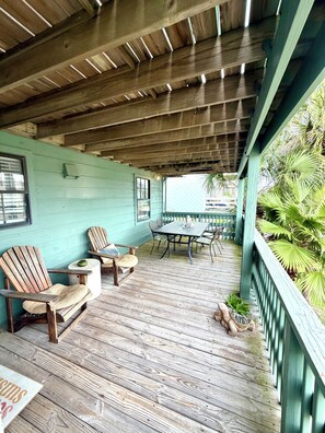 A covered outdoor area with wooden flooring, a table with chairs, and a view of greenery.