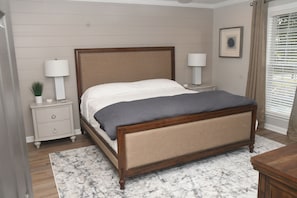 A bedroom with a wooden bed, two bedside tables with lamps, a nightstand with a plant, a marble rug, and a framed picture on the wall.