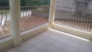 Een balkon met betegelde vloer, metalen hekwerk en uitzicht op een aangelegd gebied met een stenen muur en een gebouw op de achtergrond.