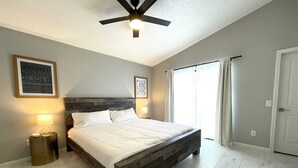 Una camera da letto con un letto con struttura in legno, una ventola a soffitto, un comodino con lampada e un'ampia finestra con tende.