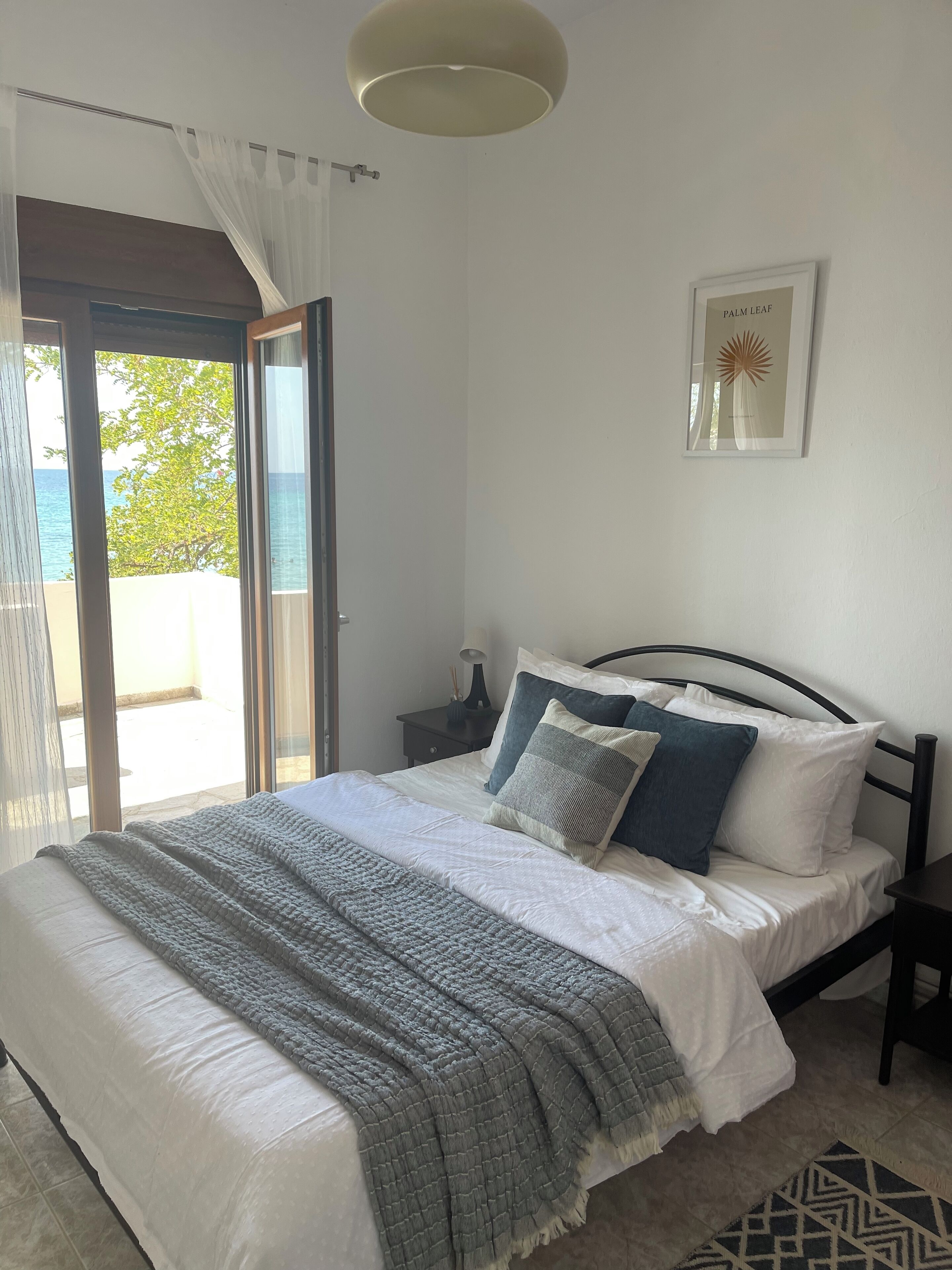 Una camera da letto con un letto, un comodino, un quadro incorniciato e vista sul mare attraverso una porta in vetro.