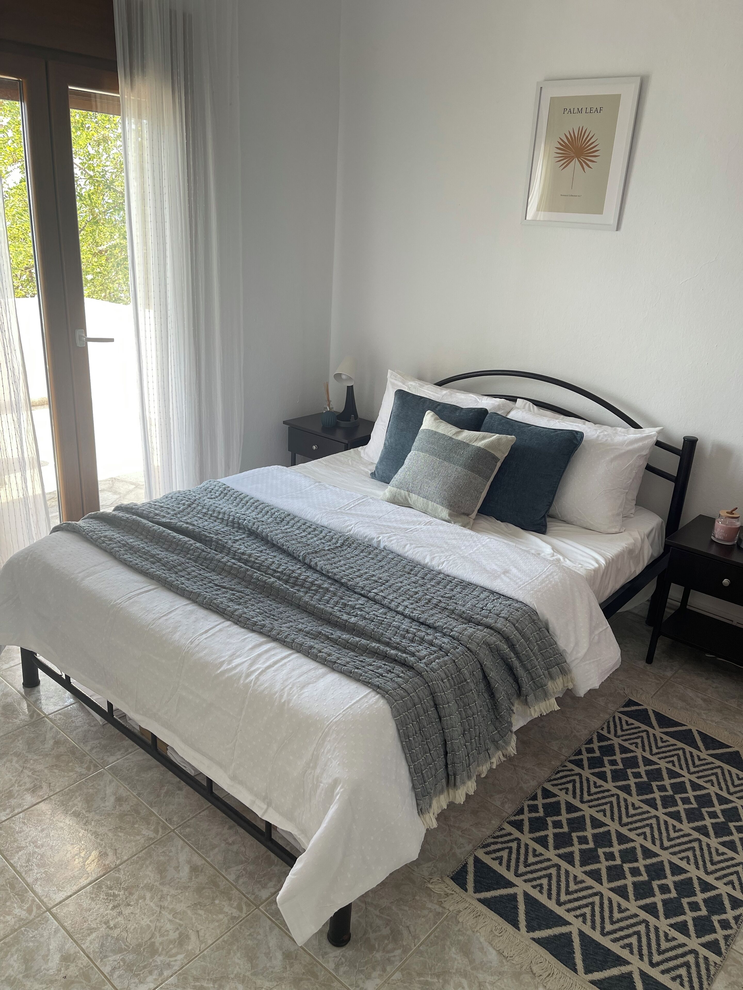 Een net opgemaakt bed met een grijs-wit quilt, een geborduurde plaid en een geometrisch vloerkleed.