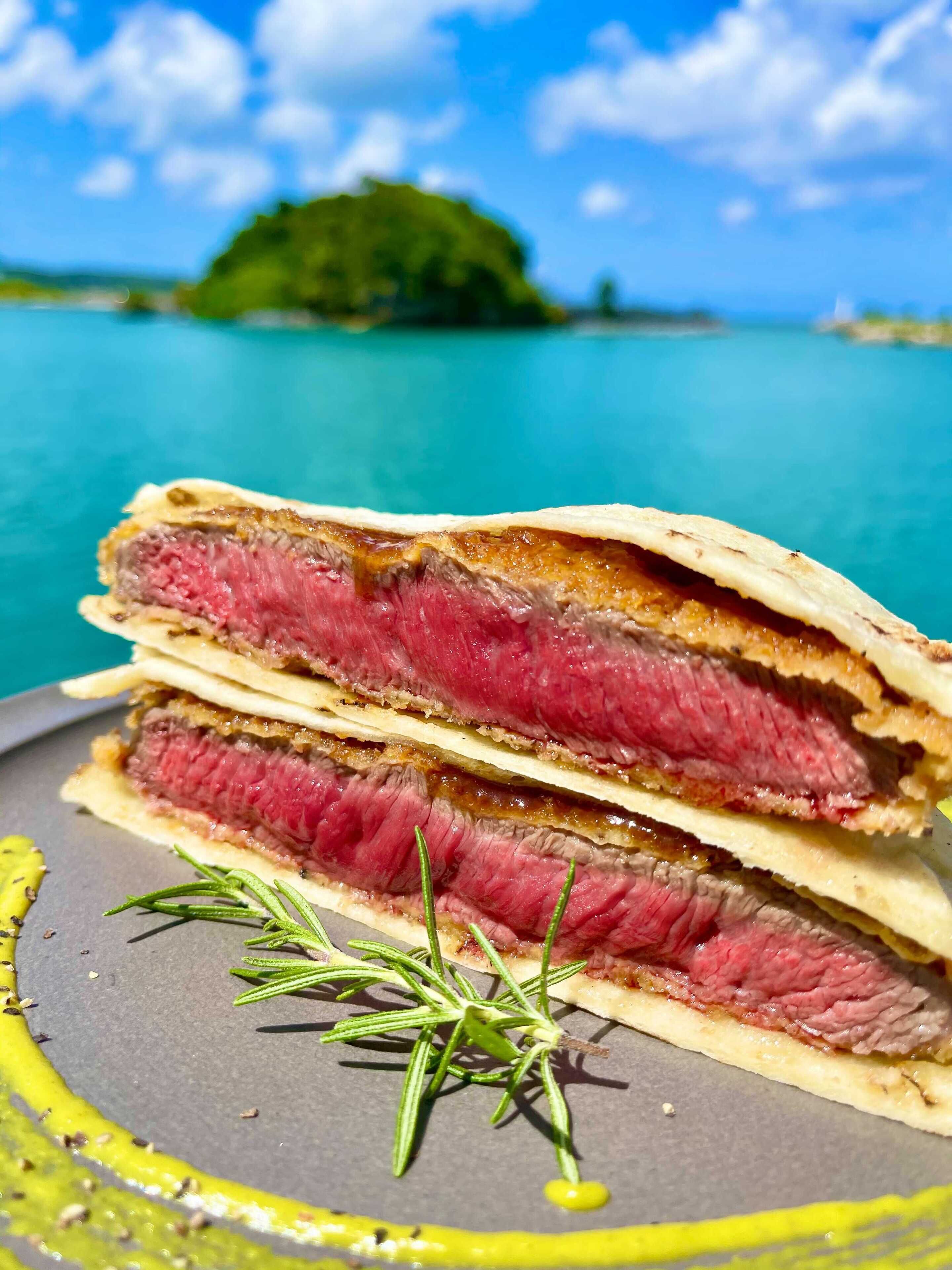 A stack of sandwiches with a sprig of rosemary on a plate, set against a scenic background of a turquoise sea and a green island.