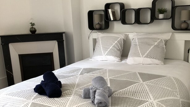 A neatly made bed with a geometric patterned bedspread, a bedside lamp, and a wall-mounted shelf with books and a plant. - Tour Eiffel cocooning apartment (Paris)