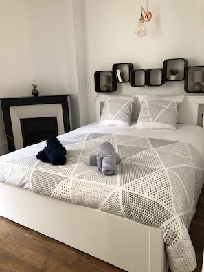 A neatly made bed with a geometric patterned bedspread, a bedside lamp, and a wall-mounted shelf with books and a plant.