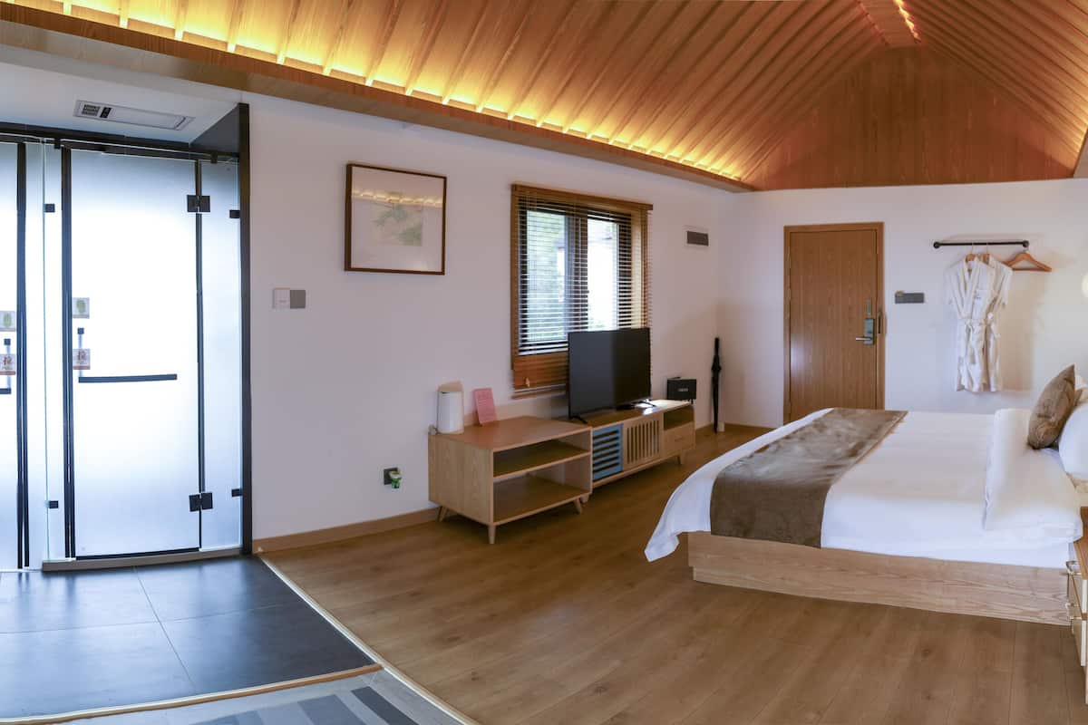 A modern hotel room with a wooden ceiling, a bed with white bedding, a TV on a wooden stand, a wooden desk with a computer, and a wardrobe.
